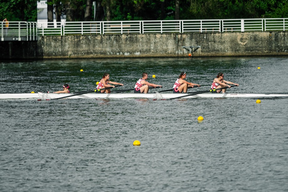 A women’s coxed four racing down a buoyed course, mid-stroke.